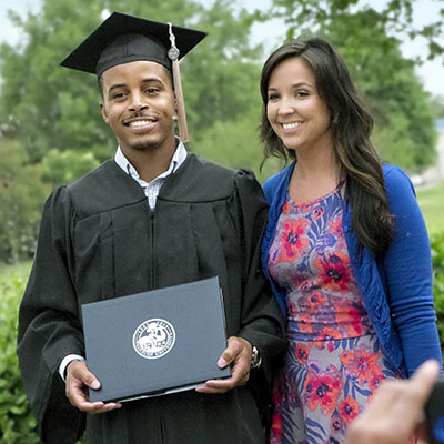 Male student in graduation robe with diploma cover, standing next to woman, having picture taken