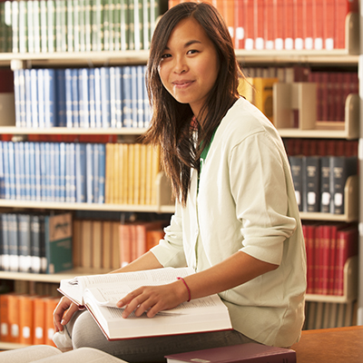 Asian female student in Mabee Library, sitting on table in front of bookshelves, looking at camera.