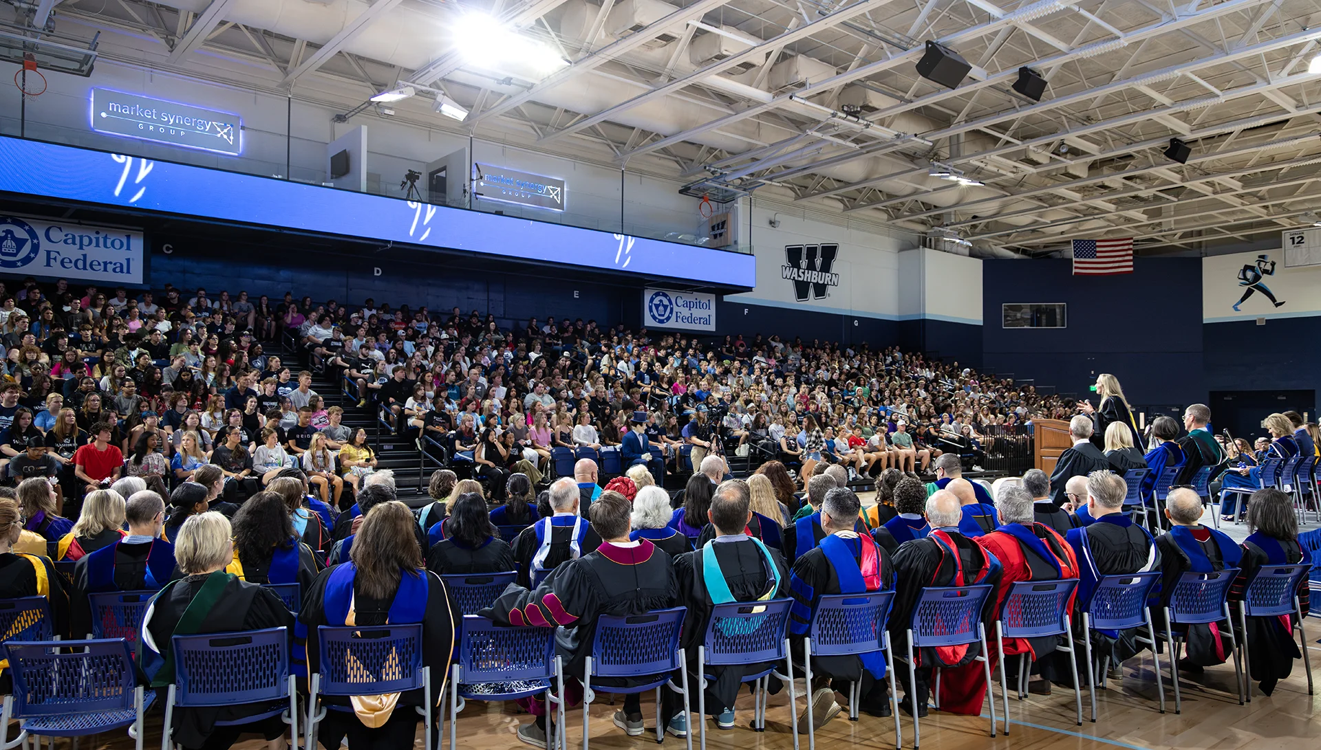 A row of faculty in graduation regalia sit in front of bleachers full of Washburn freshmen for Convocation.