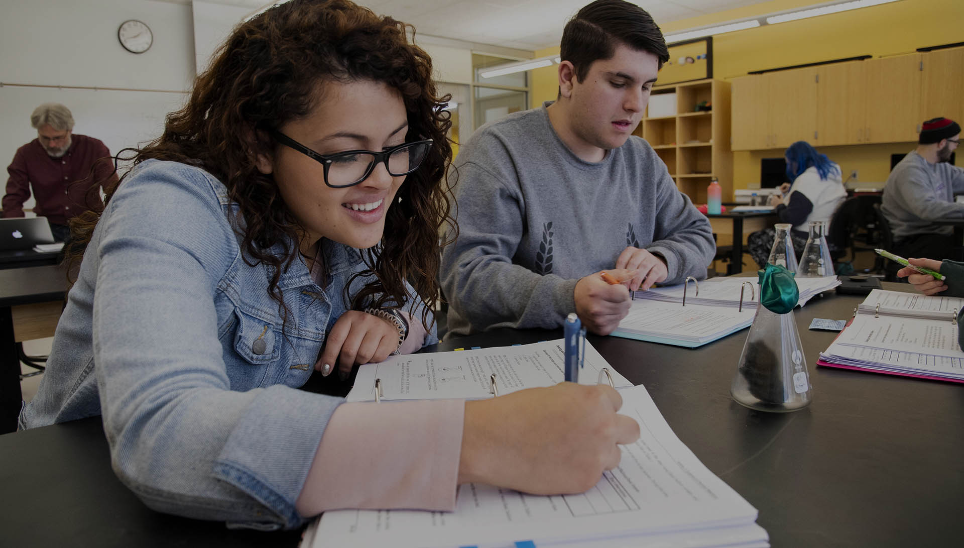 A student smiles while taking notes during a physics lab.