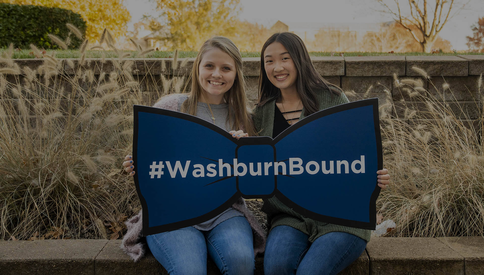 Two students smile and pose with a sign in the shape of a bow tie that says #WashburnBound.