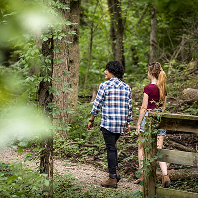 Two people walk on a trail in a forested area.