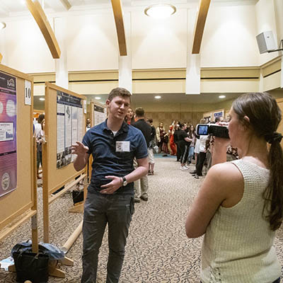 student discussing information on poster to person taking video