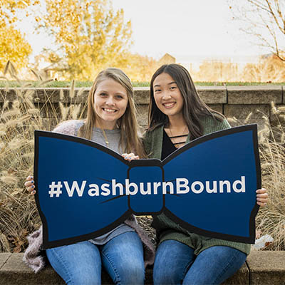 Two future students smile and pose with a giant bowtie that says #WashburnBound.