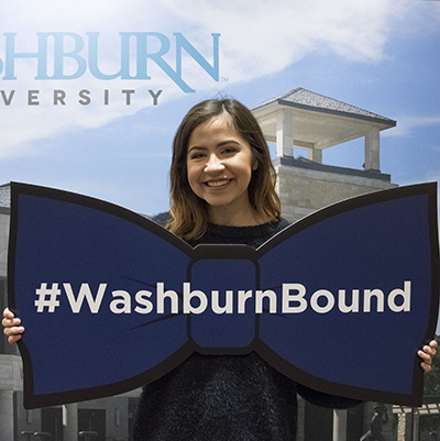 A student smiles while holding a bowtie sign that says #WashburnBound
