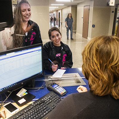 Two students smile as a business office employee helps them.
