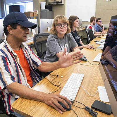 A professor gestures while sitting at a line of computers with students working on animation graphics.