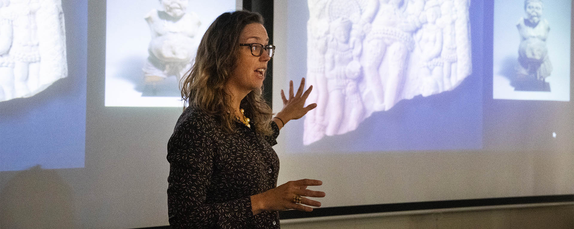 A professor gestures while standing in front of a presentation that includes photos of art pieces.