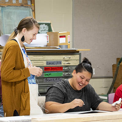 A student wearing an apron smiles while talking to another student sketching on ink.