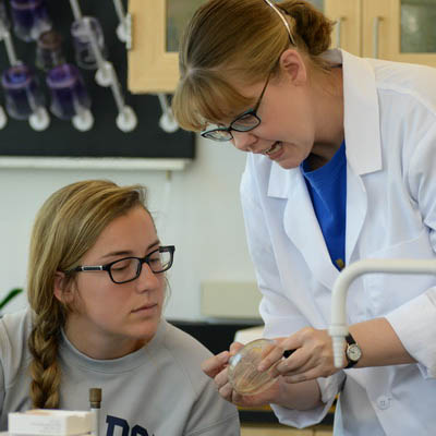 A professor wearing a lab coat explains something to a student at a microscope.