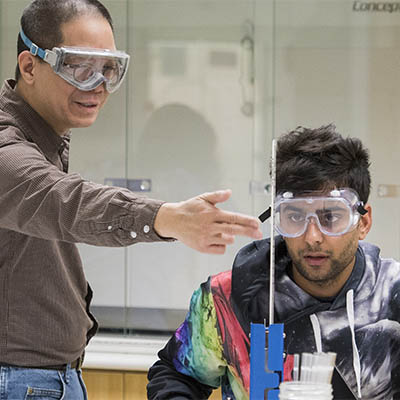 A professor points to a test tube while talking with a student in a chemistry lab.