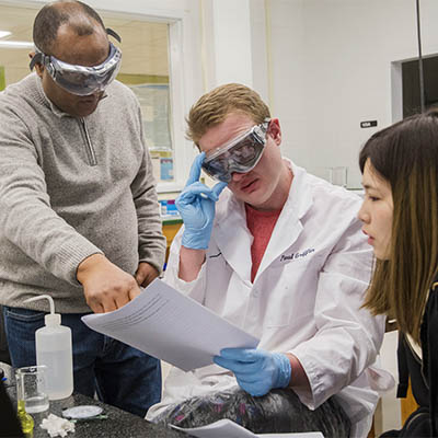 A chemistry professor points out something on a page while a student adjusts his goggles.