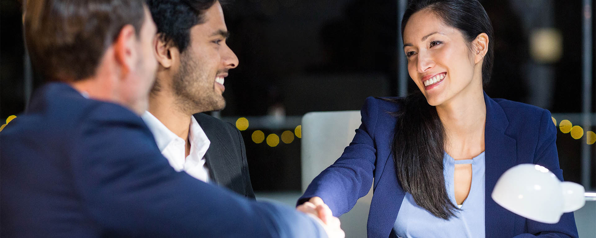 A woman wearing business attire shakes hands with a colleague at a table.