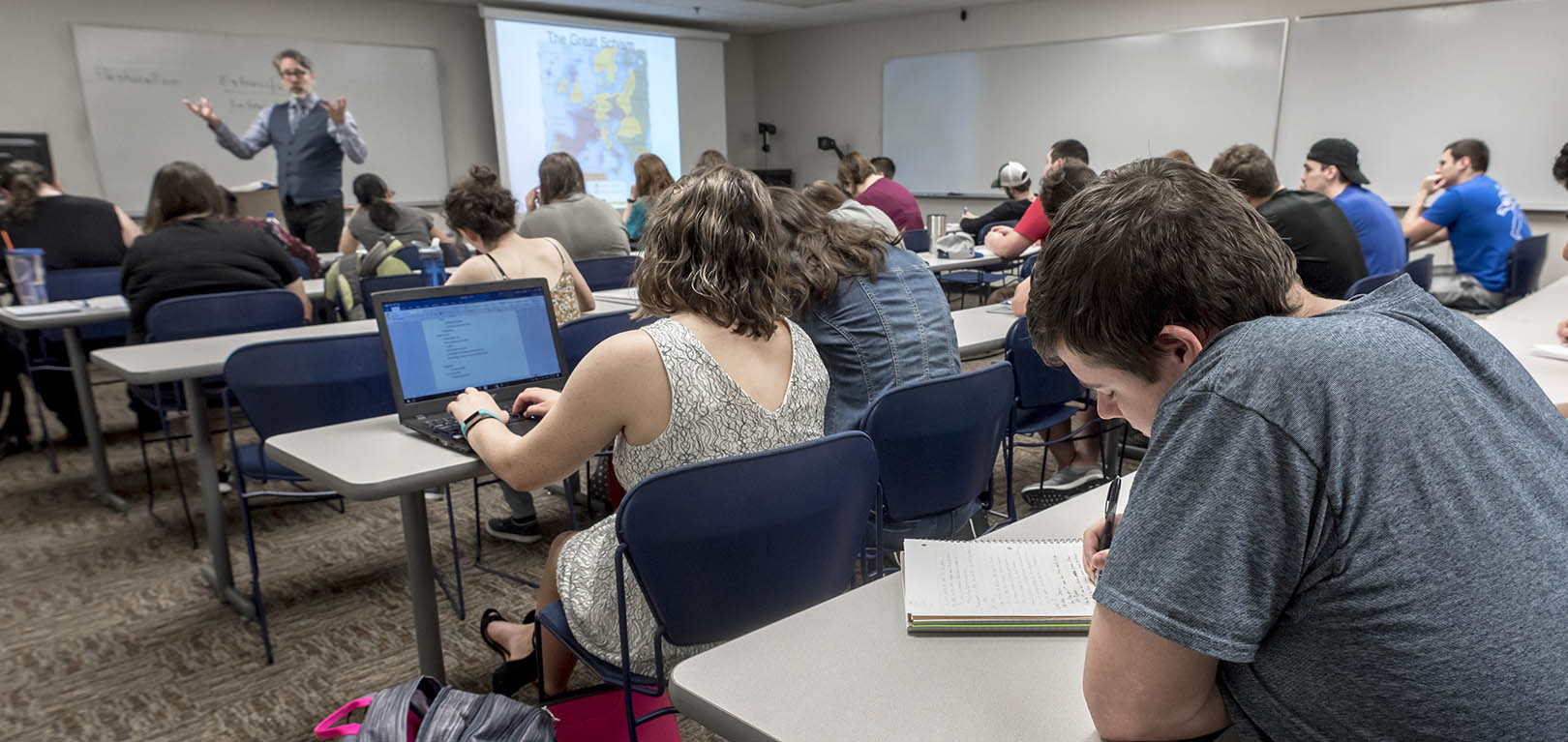 A student takes notes while a professor teaches at the front of the room.