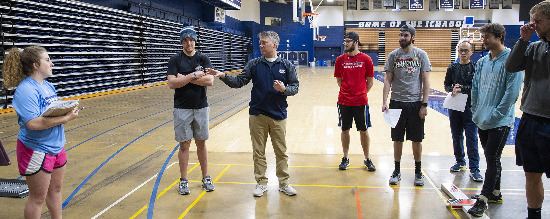 Students gather around a professor in a gym as he explains the day activities.