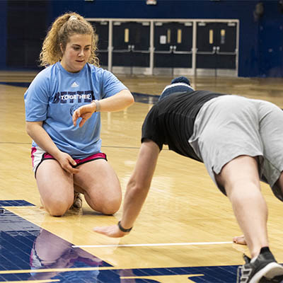 A student watches her stop watch as another student does pushups in a gym.