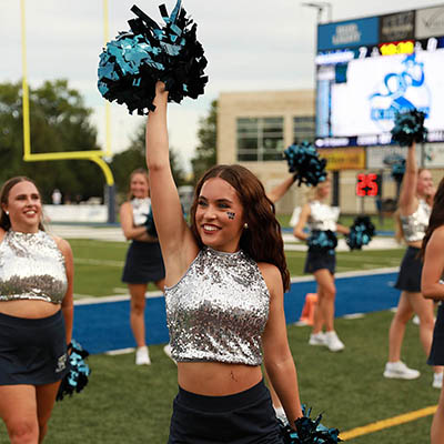 A dancing blue in silver top and black skirt cheers with other blues on football field