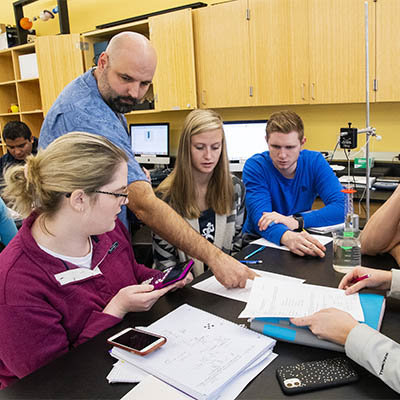 A professor points something out in a textbook to a group of physics students in lab.