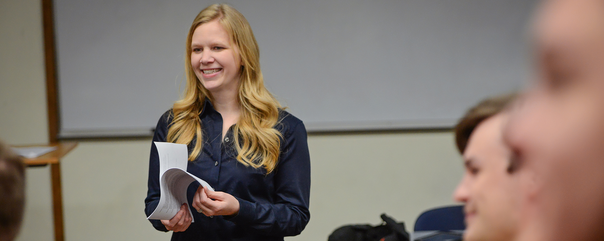Blonde woman smiles while holding papers in front of classroom