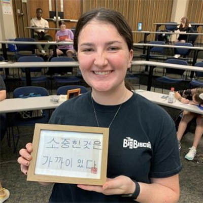 A student smiles holding a framed piece with Korean words on it.