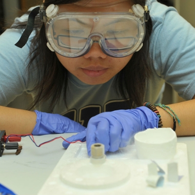 A forensic chemistry student wearing goggles and gloves stares intently at a sample.