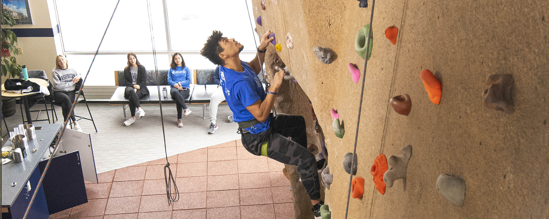 A student climbing the rock wall while others watch from below.