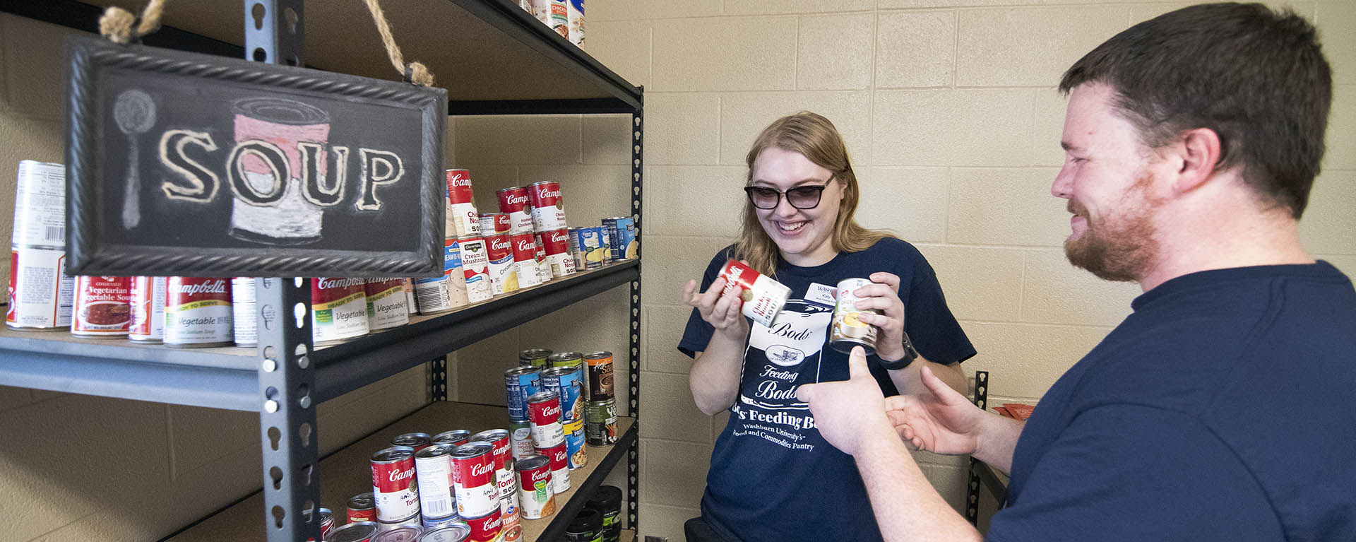 Two students smile while stocking the pantry shelves with cans of soup.