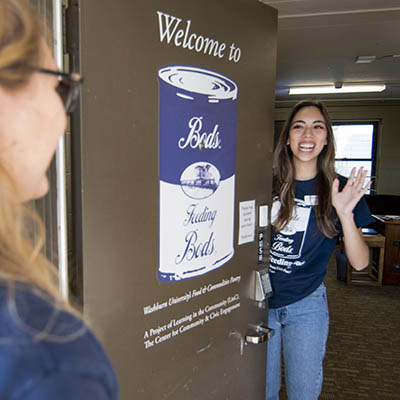 A student smiles and waves while welcoming someone into the pantry.