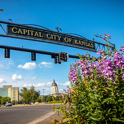 Flowers bloom as the Capital City of Topeka sign spans overhead with the capitol in the background.