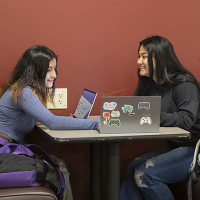 Two students talk while working on their laptops.