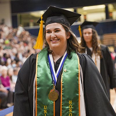 A student with an honors stole walks smiles at graduation.