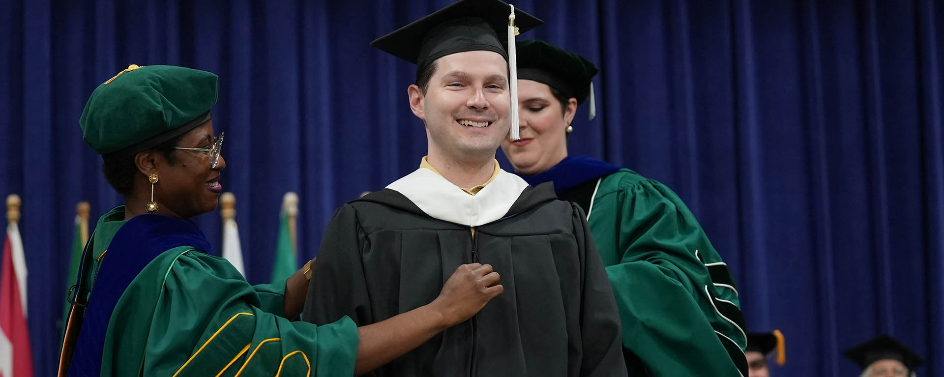 A graduate student smiles while receiving their hood at graduation.