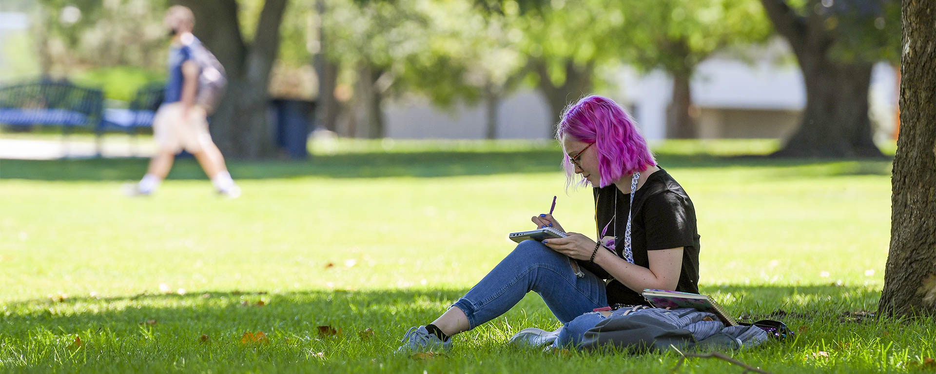 A student writes in a notebook while sitting in the grass under a tree on campus.