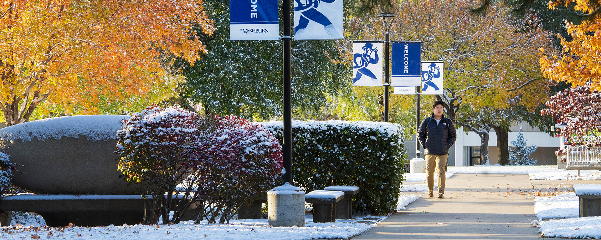 Fall colored trees glow in the morning light while snow dusts the ground as a student walks to class on campus.