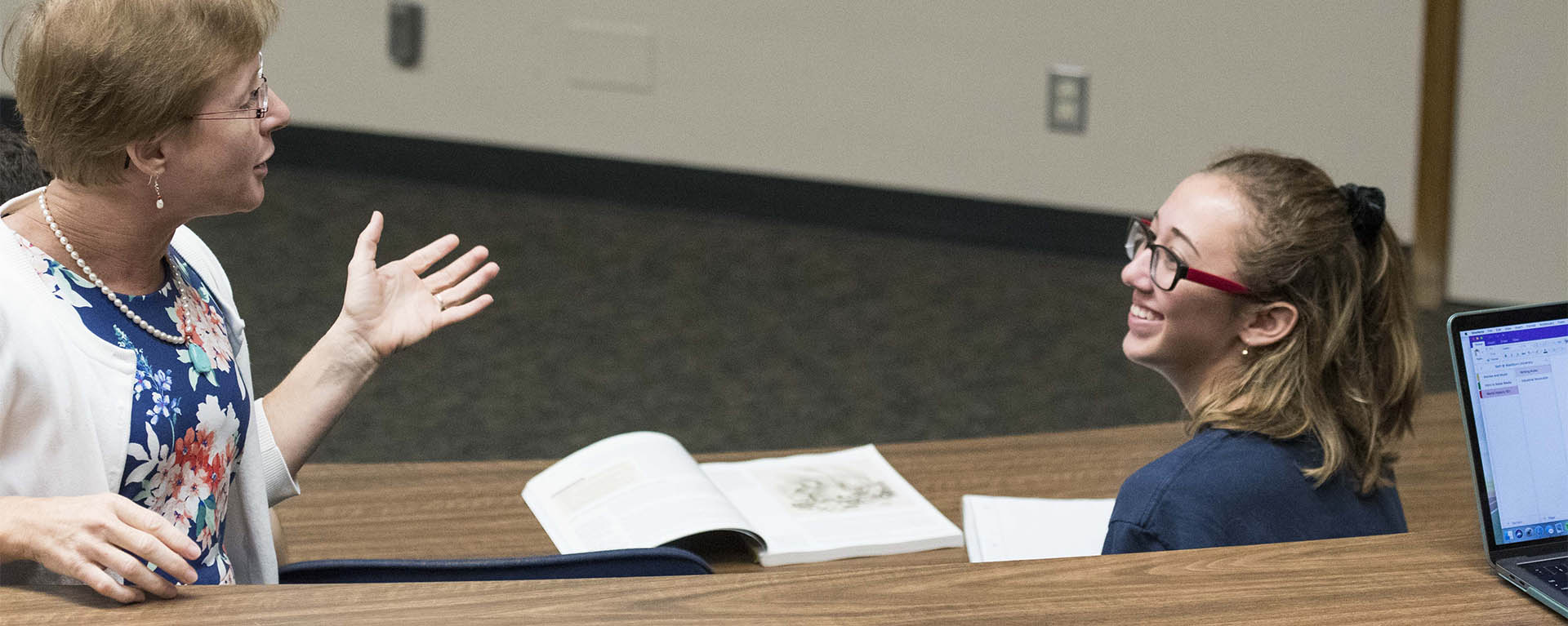 A professor gestures while talking to a smiling student during a class lecture.