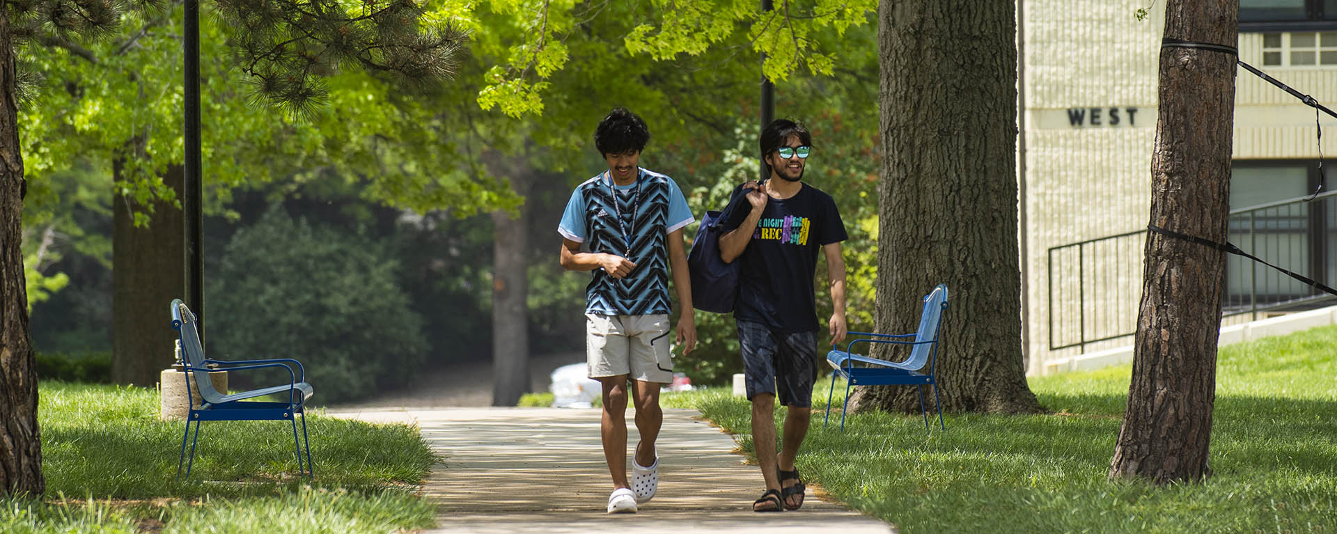 Two students smile and chat while carrying laundry as they walk across campus on an early summer day.
