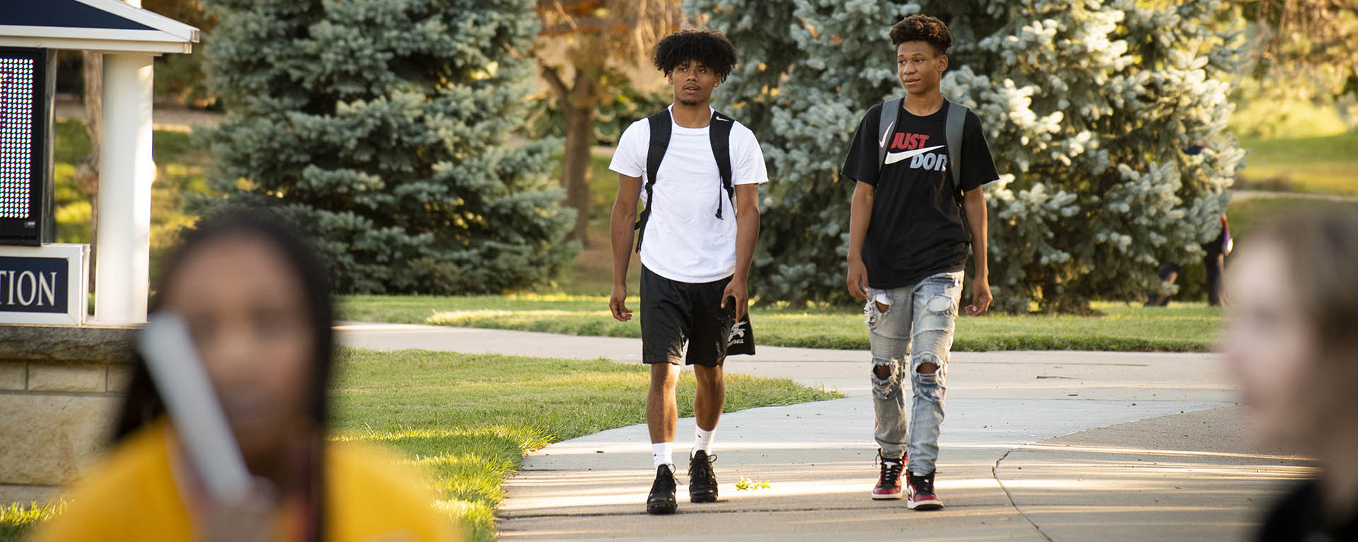 Two students smile and talk while carrying their backpacks as they walk through campus on an early fall day.