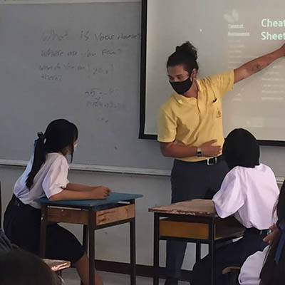 A student wearing a mask gestures to the board while teaching a class of students.